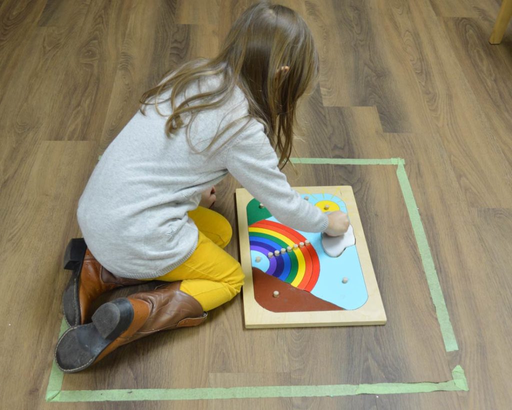 A child sitting in a taped off box on the floor and working on a rainbow puzzle.